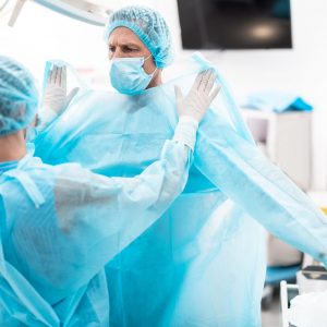 Medical workers. Portrait of nurse in sterile gloves putting on blue surgical gown on doctor