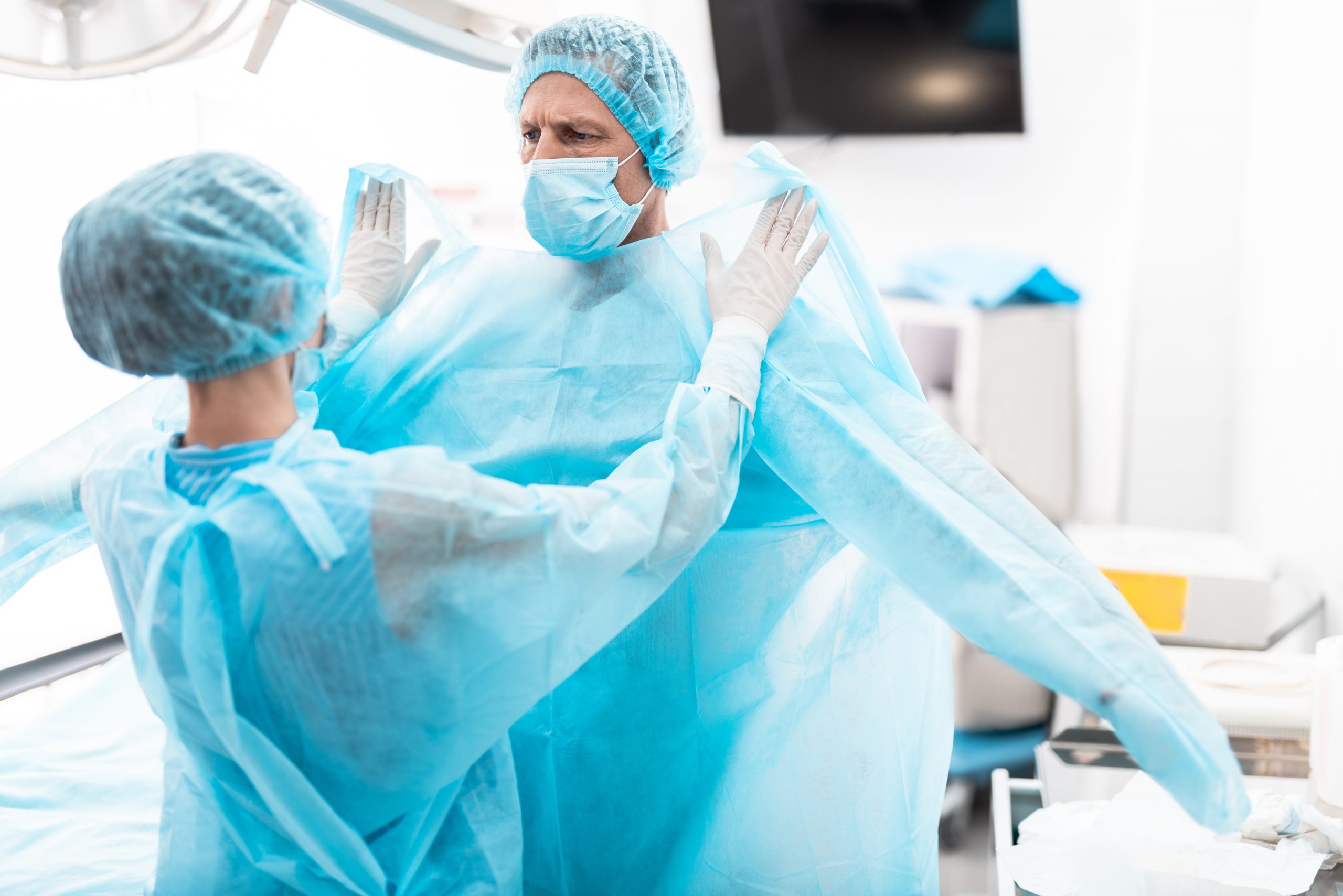 Medical workers. Portrait of nurse in sterile gloves putting on blue surgical gown on doctor